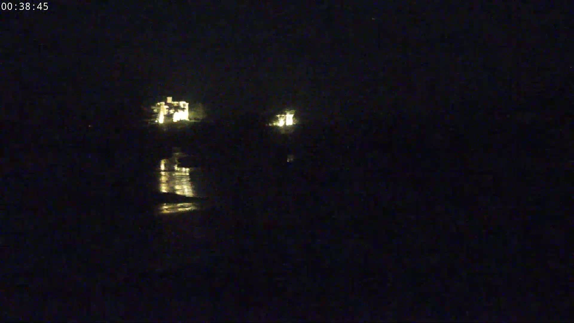 At night, two illuminated structures are visible across a dark body of water, whose surface reflects the lights.