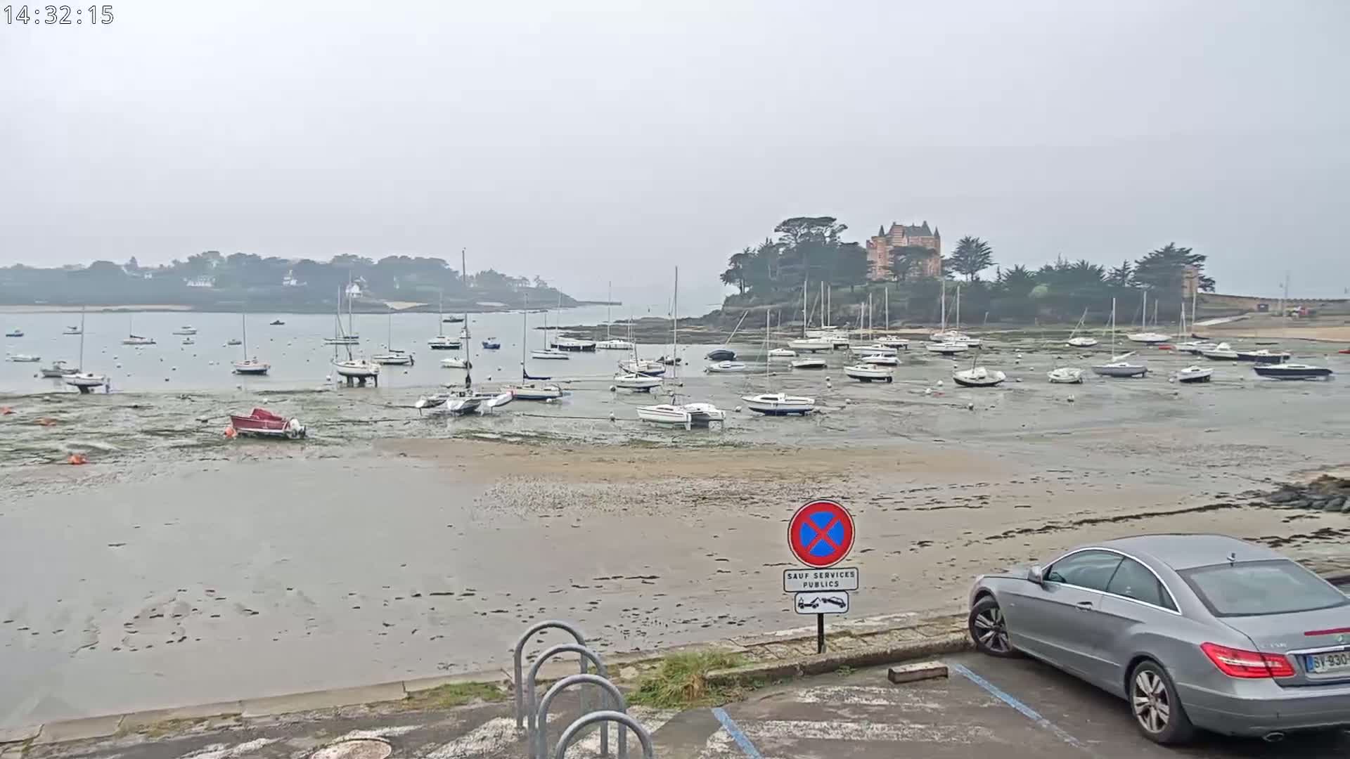 Numerous sailboats are grounded on a wide, muddy, low-tide beach in front of a large chateau-like building on a cloudy day.