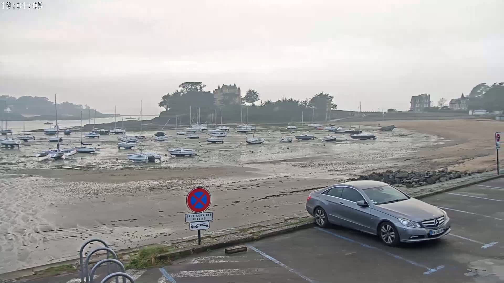 A gray car is parked in a lot overlooking a sandy beach where numerous sailboats are grounded at low tide under an overcast sky.