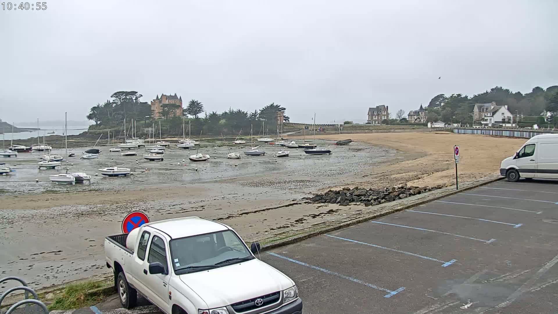 A low-tide beach scene shows numerous sailboats grounded on a sandy expanse, near a shoreline with buildings and a white pickup truck parked in a lot.
