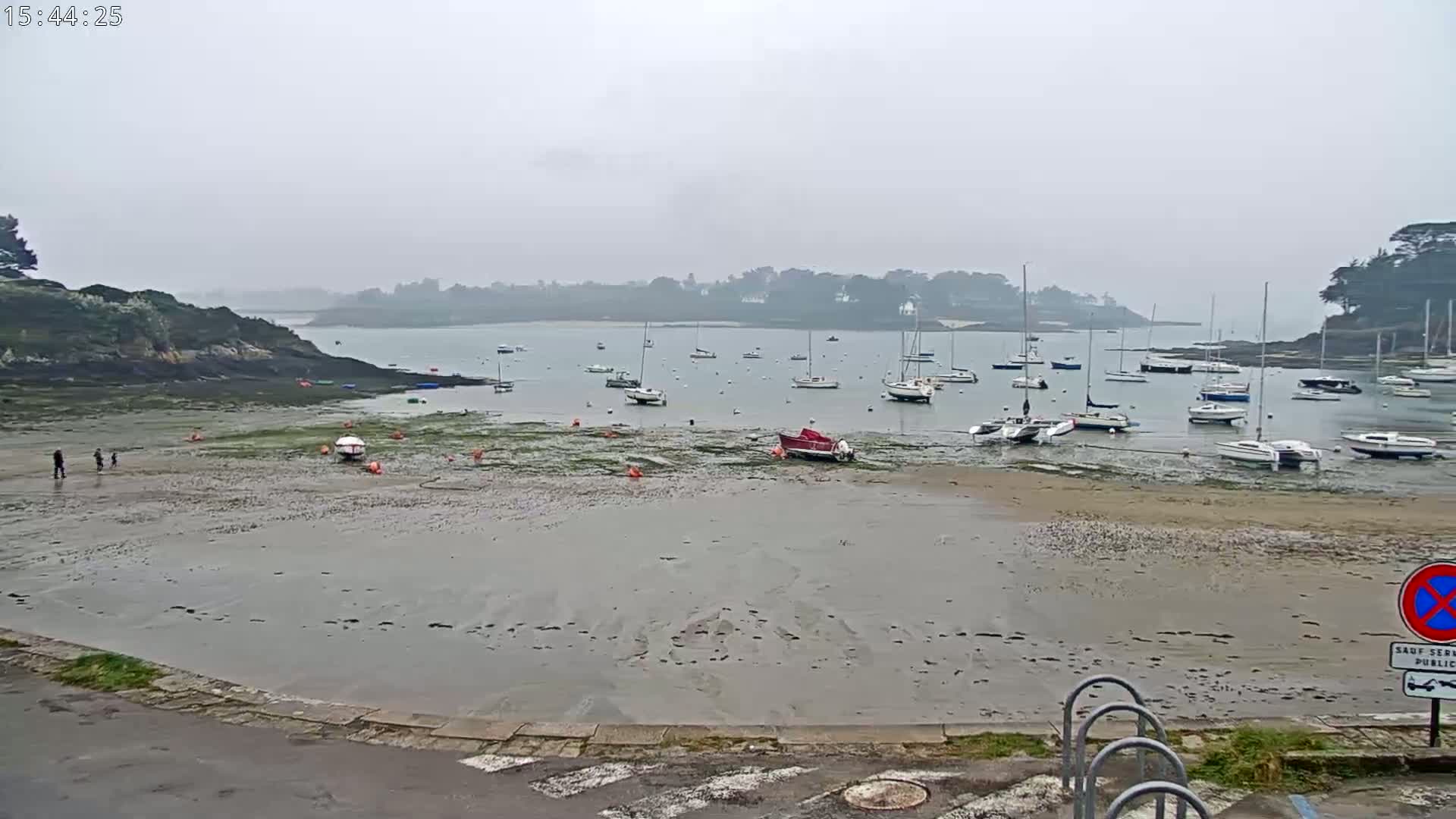 A mostly overcast day reveals a calm bay with numerous sailboats at rest in shallow water, a sandy shore, and a few people walking on the exposed seabed.