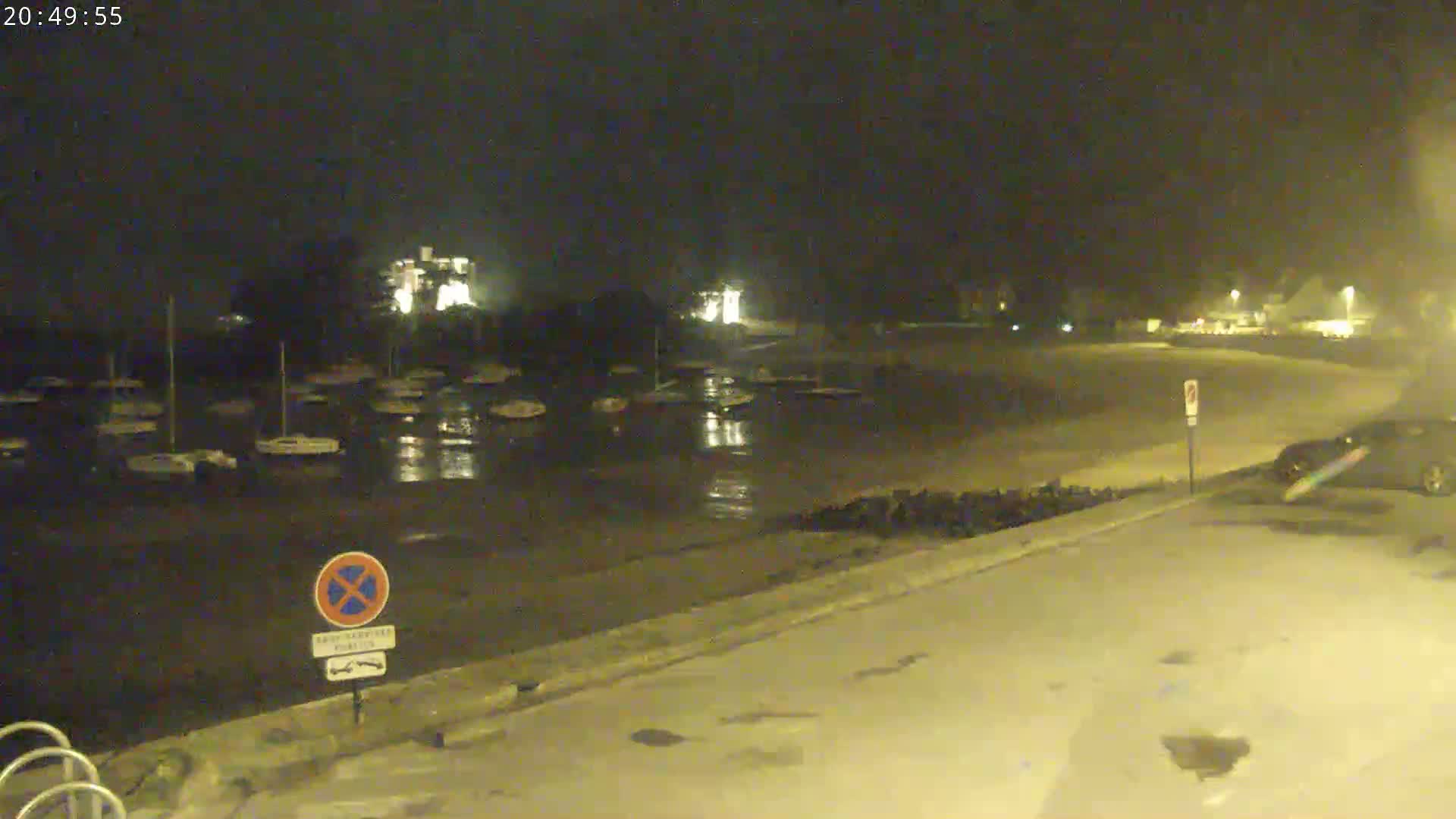 At night, a wet, empty parking lot next to a harbor filled with docked sailboats is illuminated by artificial lights.