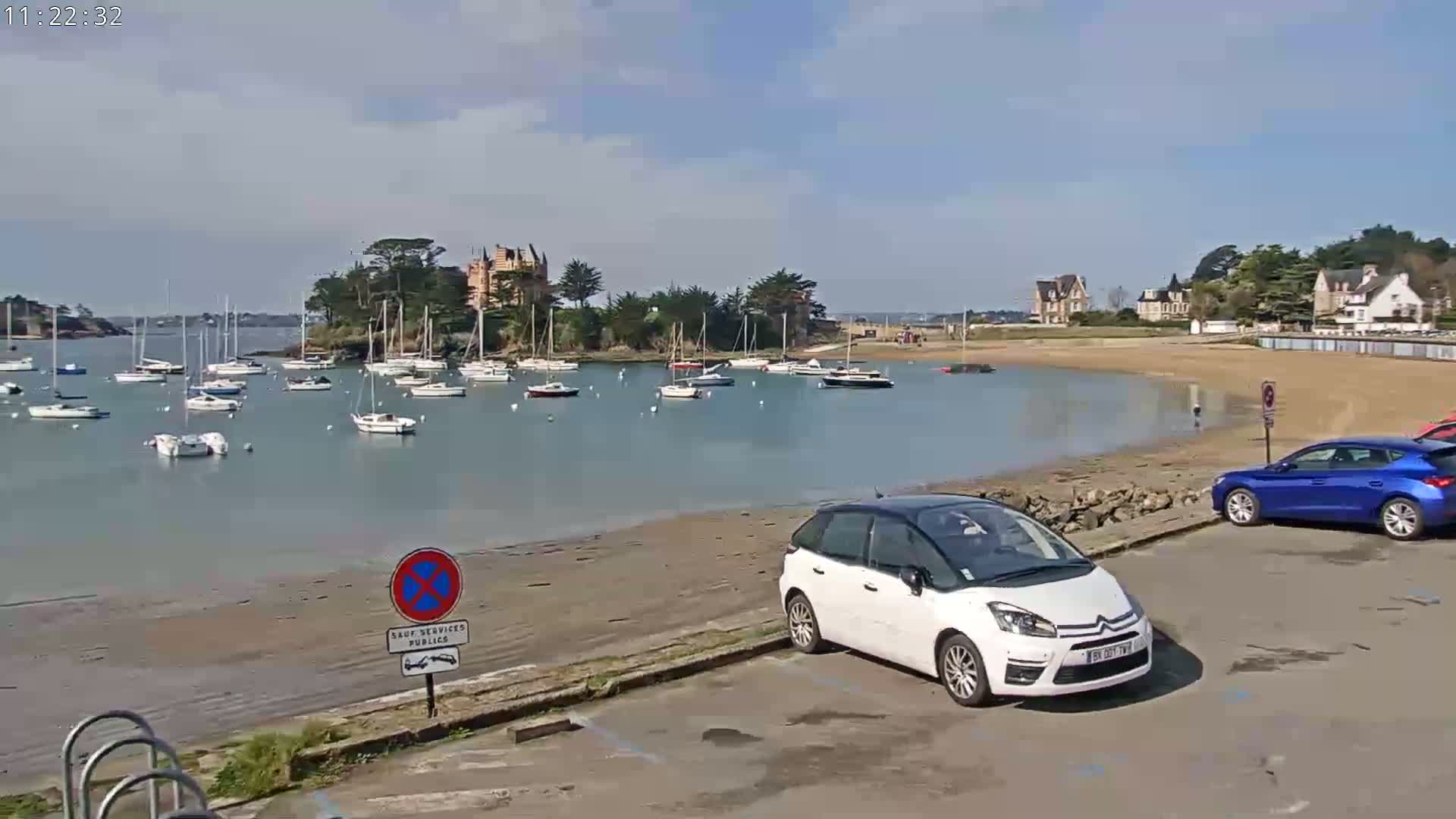 A calm bay with numerous sailboats, a sandy beach, and several cars parked near buildings on a sunny day.