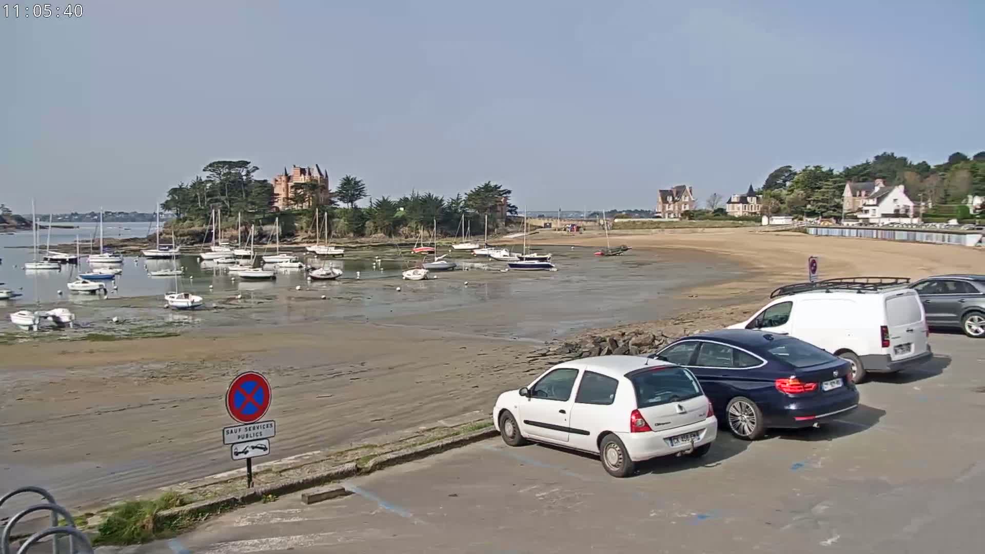 A sandy beach with several sailboats in the water and cars parked in a lot, under sunny skies.