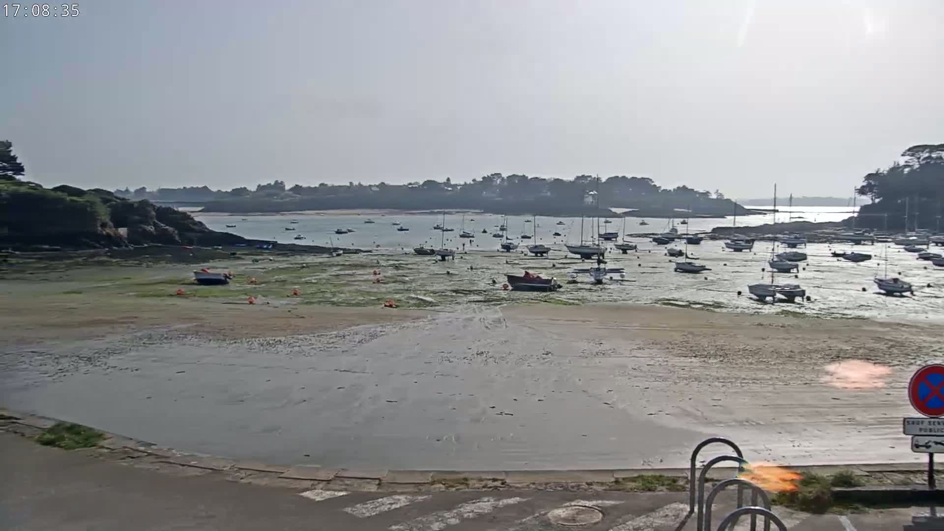 A mostly sunny day reveals a sandy inlet with numerous boats at rest on shallow, seaweed-covered waters, backed by a calm bay and distant tree-lined hills.