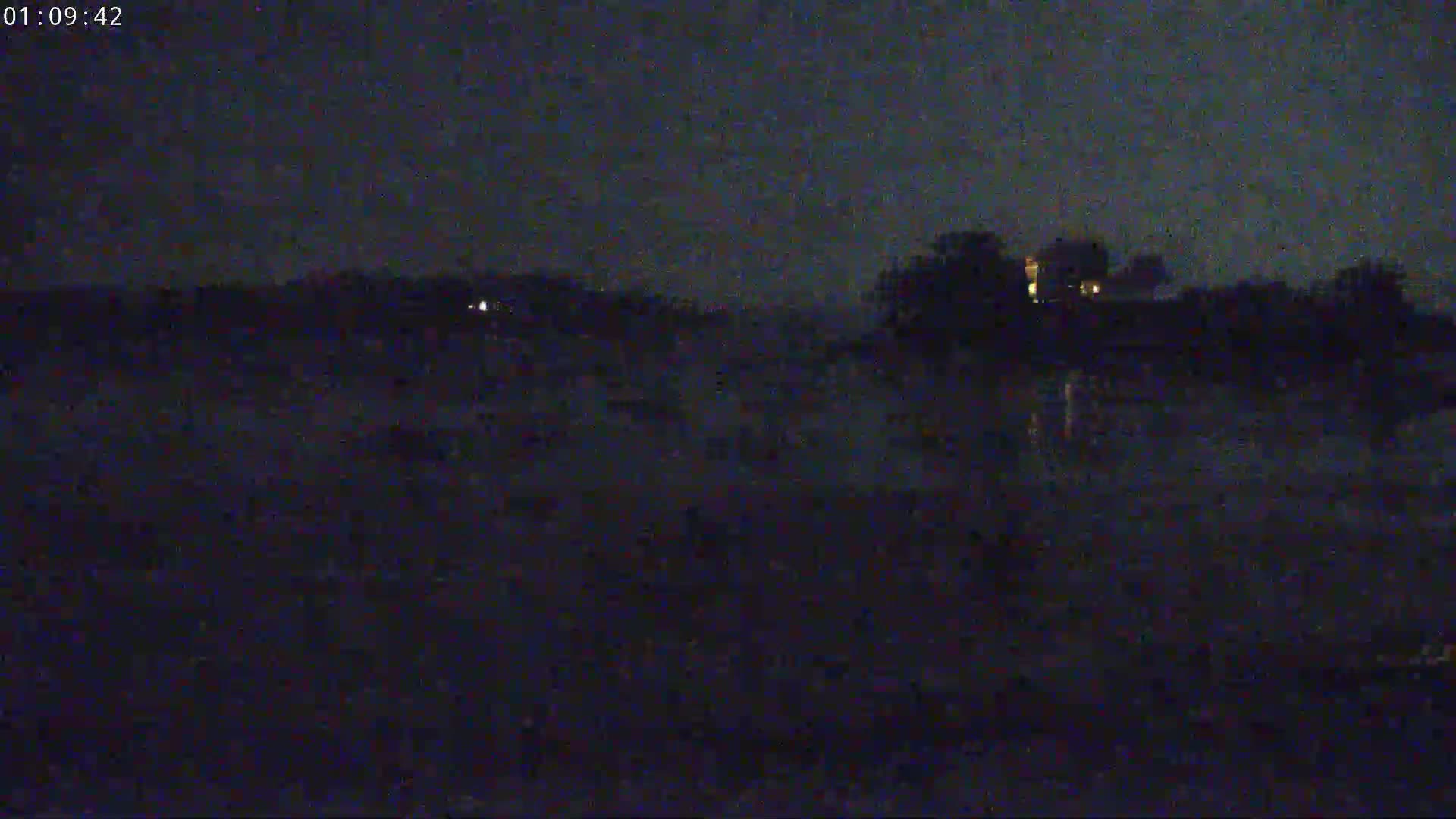 A dark, nighttime scene shows a dimly lit building and trees reflected on still water under a mostly clear sky.