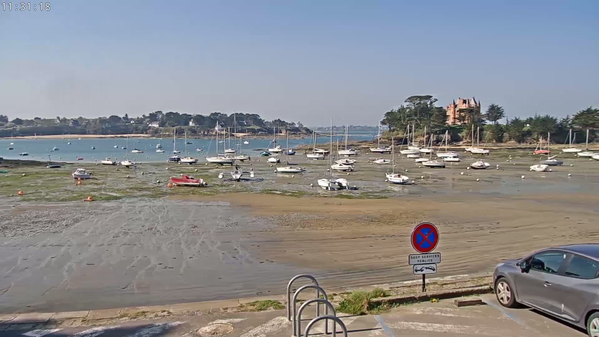 Many sailboats are resting on a wide, sandy, low-tide beach near a calm body of water and a chateau, under a clear, sunny sky.