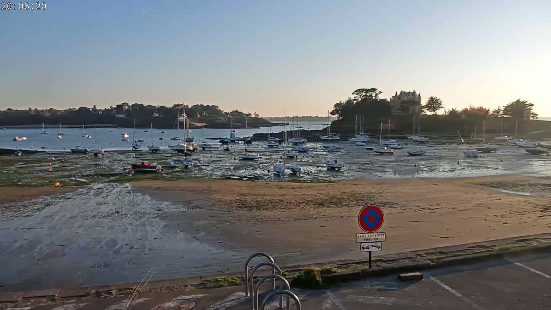 Many sailboats are moored in a calm harbor at low tide, with a sandy shore in the foreground and a building on a hill in the background under a clear sky.