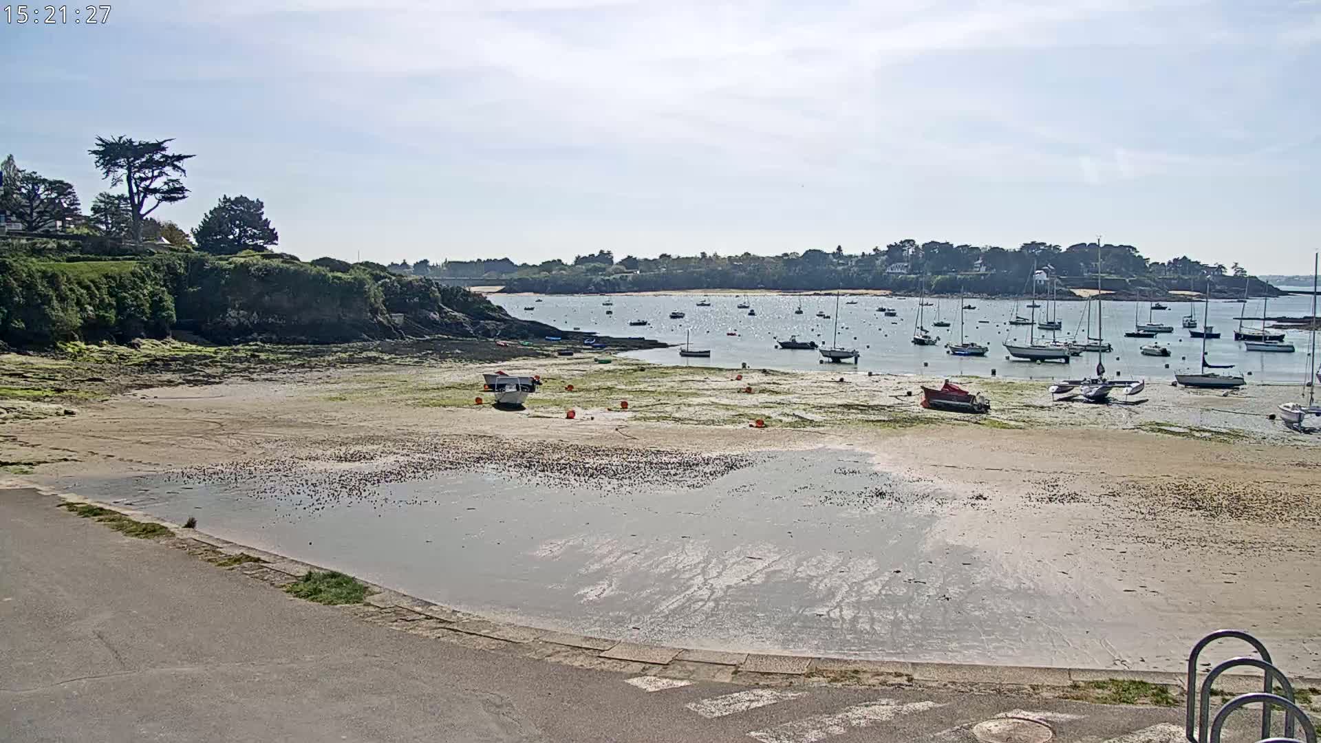 A sandy beach with a calm body of water, numerous sailboats, and a small rocky outcrop under a mostly sunny sky.