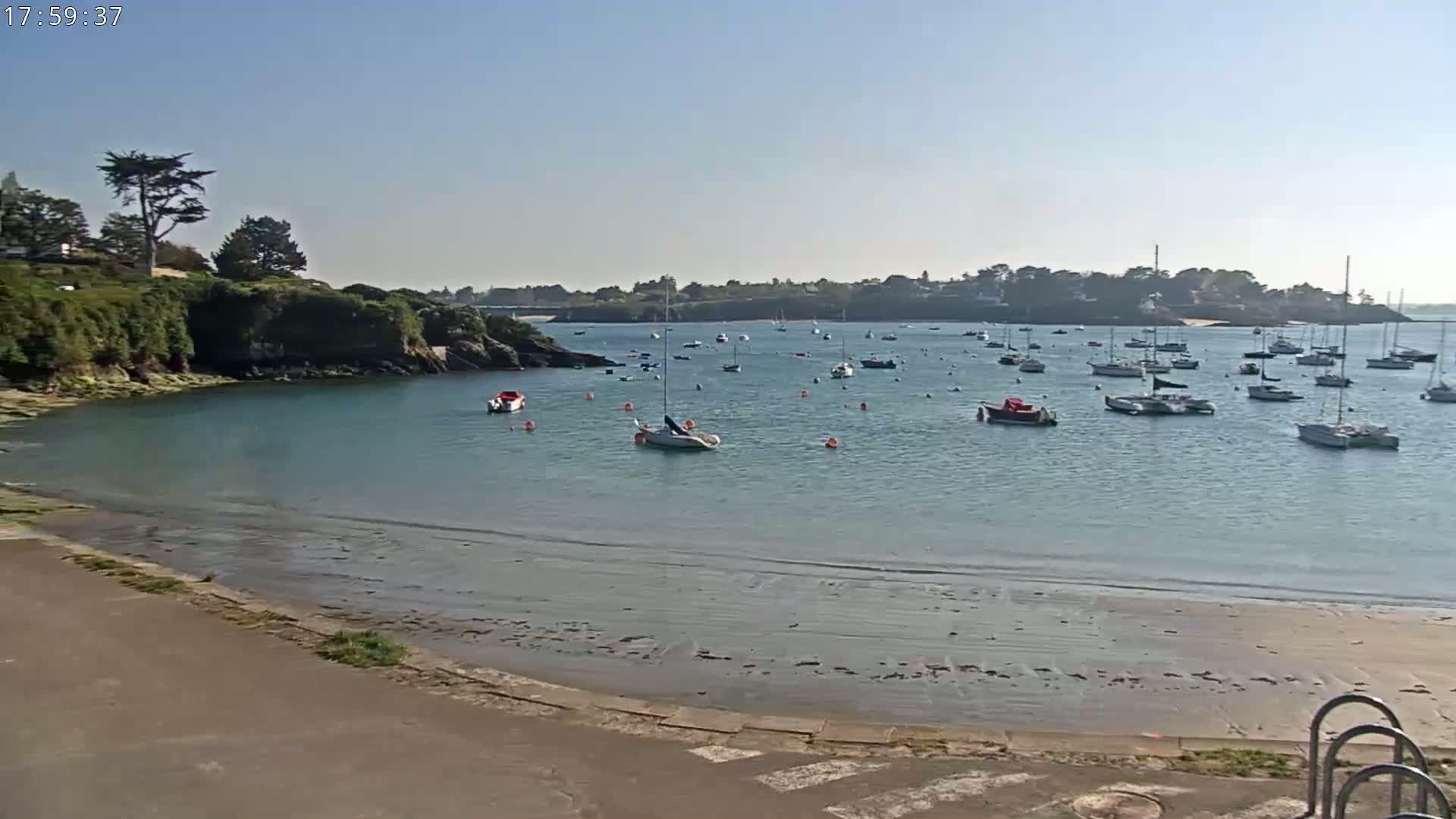 A sandy beach and calm bay filled with numerous sailboats and motorboats under a clear, sunny sky.