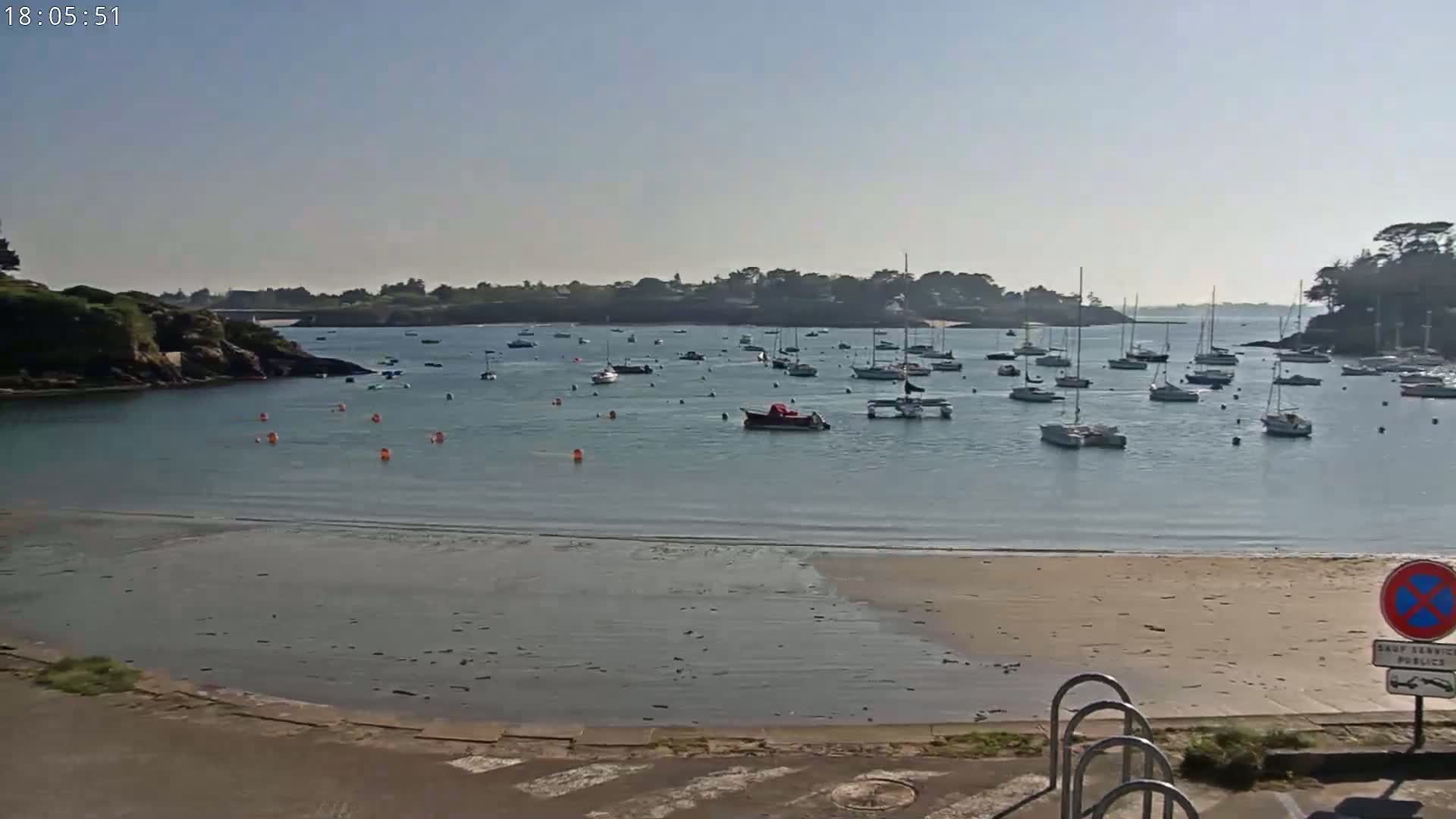 A calm bay with numerous boats at anchor is seen from a sandy shore on a sunny day.