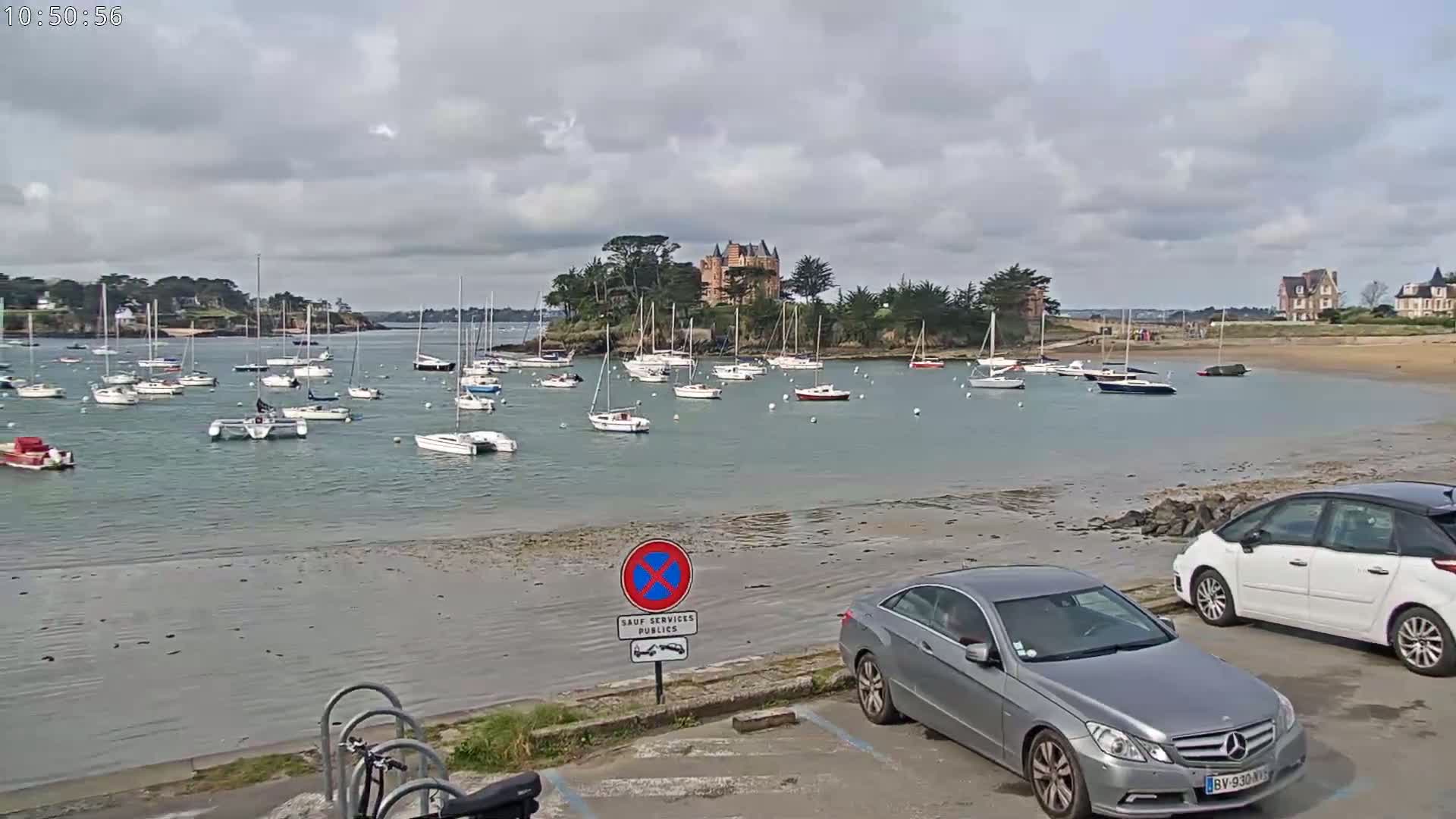 A calm bay with numerous sailboats at anchor, a château on a small island in the background, and overcast skies.