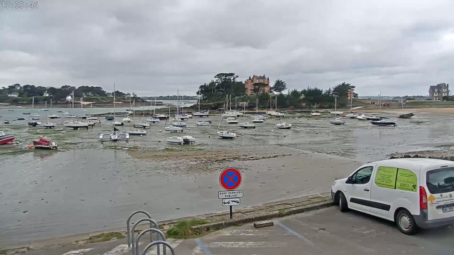 Numerous sailboats are grounded on a tidal flat near a shoreline with a chateau-like building visible in the background under an overcast sky.