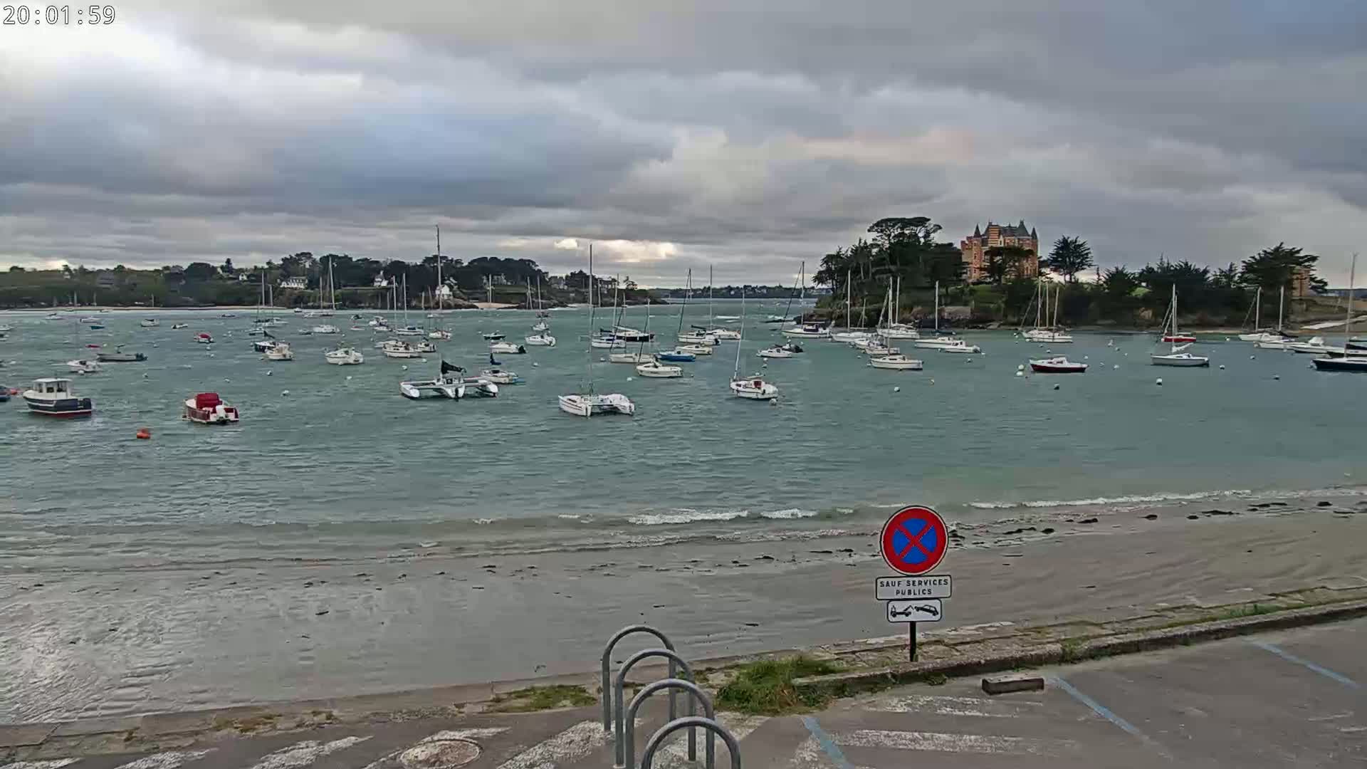 Numerous sailboats are moored in a calm bay under a cloudy sky, with a shoreline in the foreground and a large building visible on a hill in the distance.