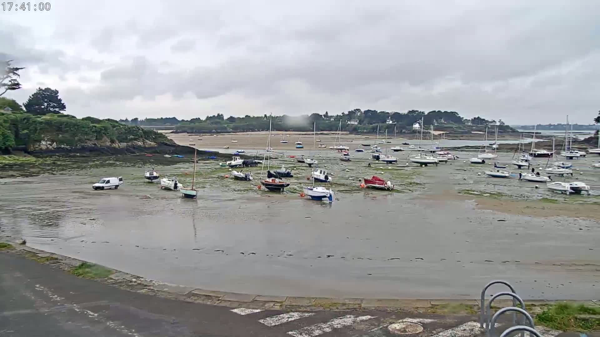 Under an overcast sky, numerous boats sit in shallow, muddy water at low tide near a small, rocky shoreline.