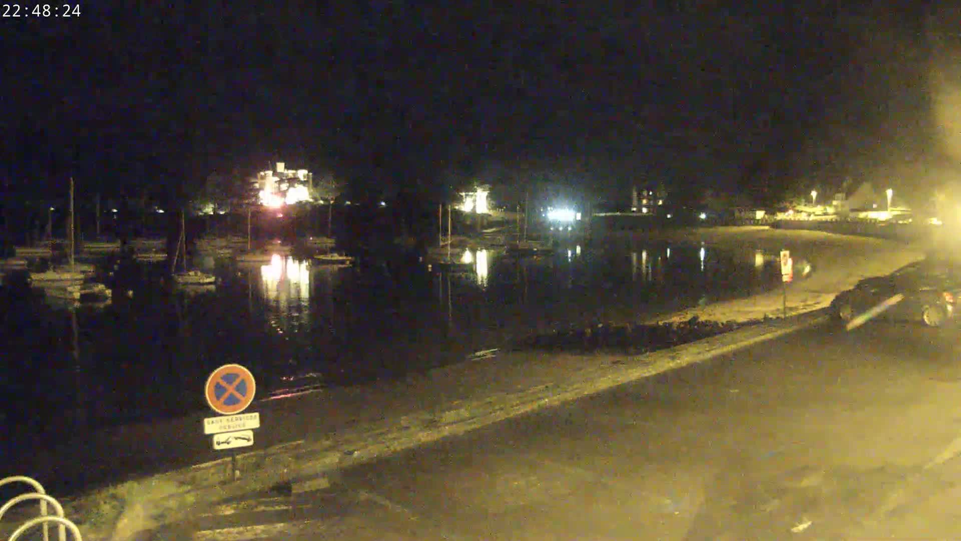 A nighttime view of a calm body of water with boats docked along the shore, illuminated buildings in the background, and a street next to the water.