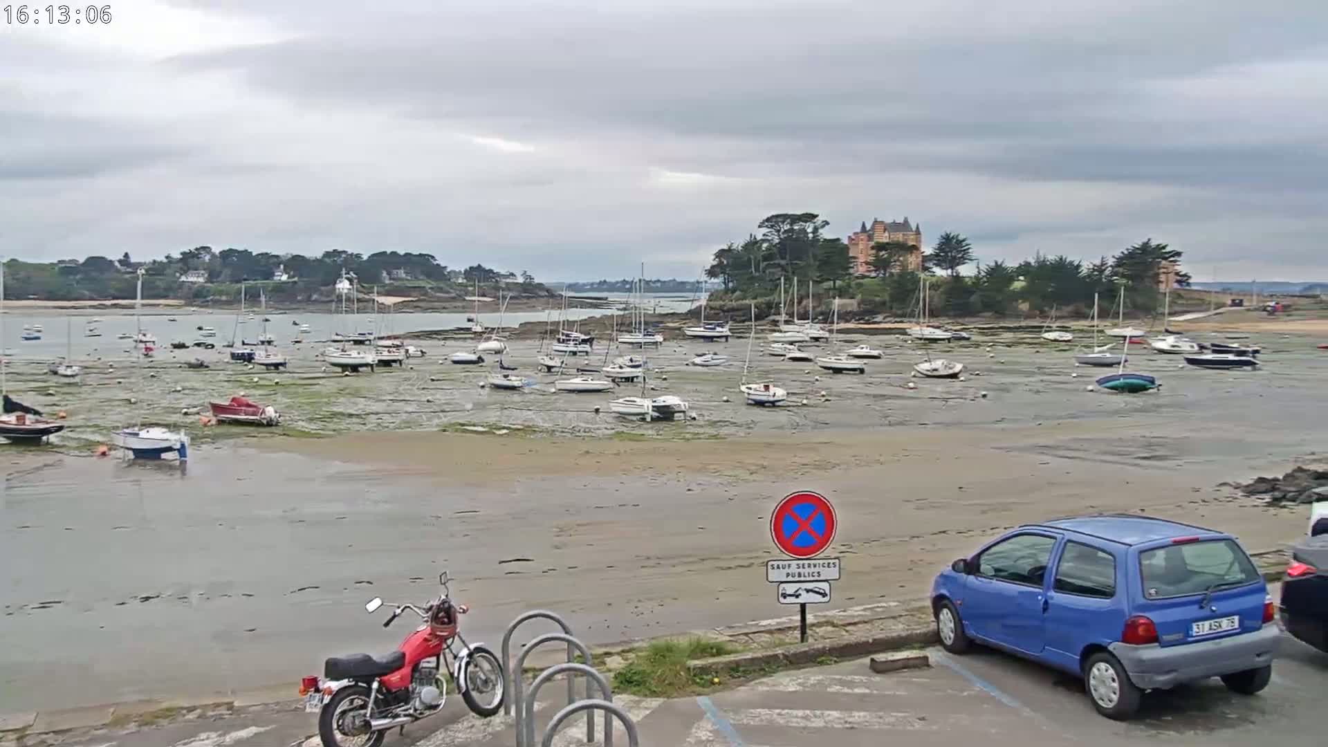 Under a cloudy sky, numerous sailboats are grounded on a tidal flat, near a small town with a large chateau visible in the distance, a motorcycle and a small blue car parked in the foreground.