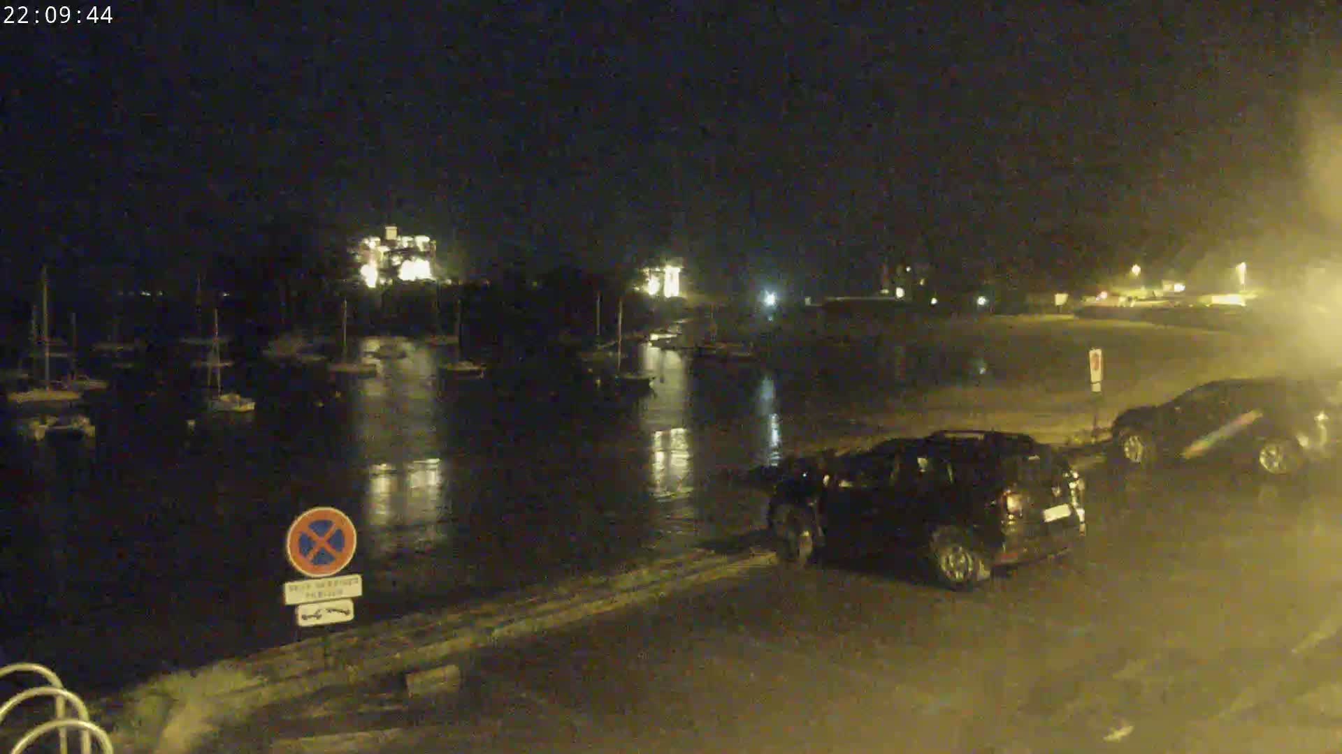 At night, a calm body of water reflects lights from buildings and boats docked along its shore, with two cars parked nearby.