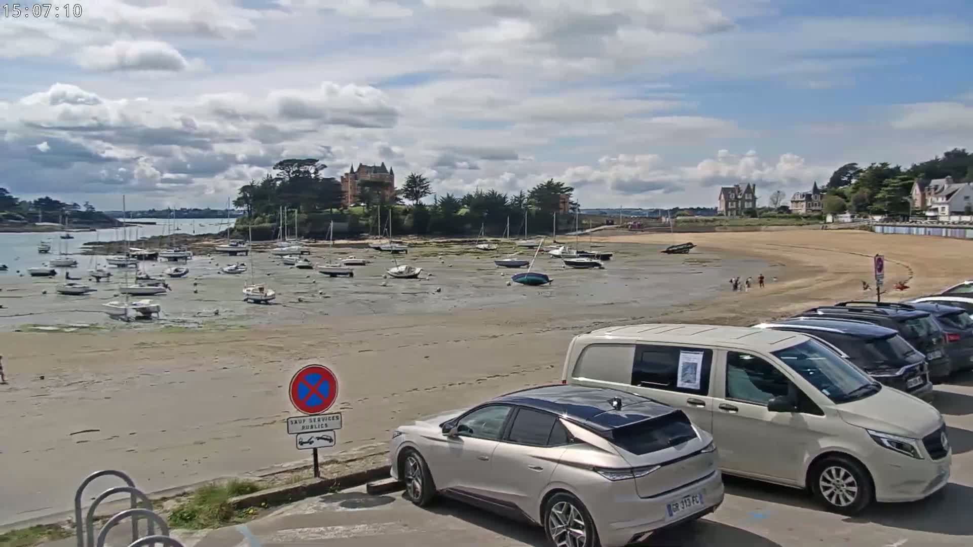 A sandy beach with numerous boats resting on exposed mudflats at low tide, under a partly cloudy sky, is overlooked by buildings and cars parked along the shore.