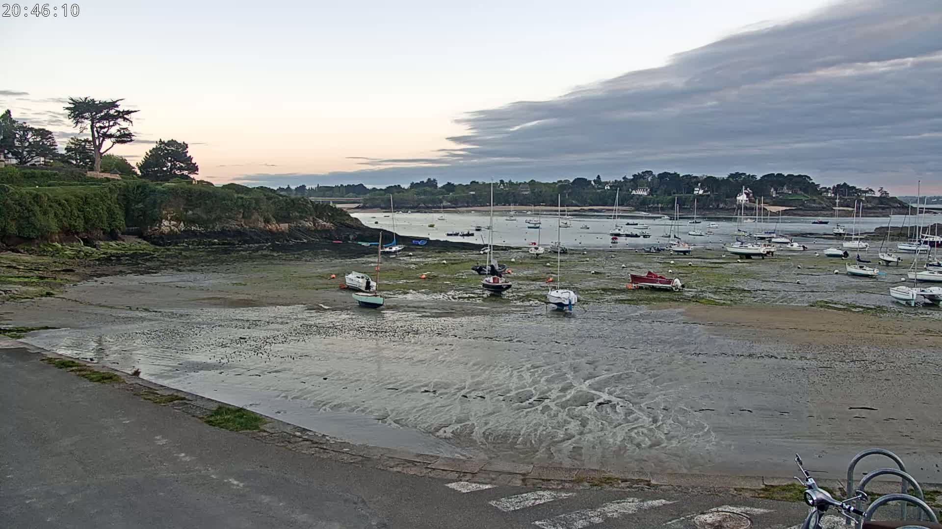 Many sailboats are grounded on a tidal flat in a calm bay under a cloudy twilight sky.