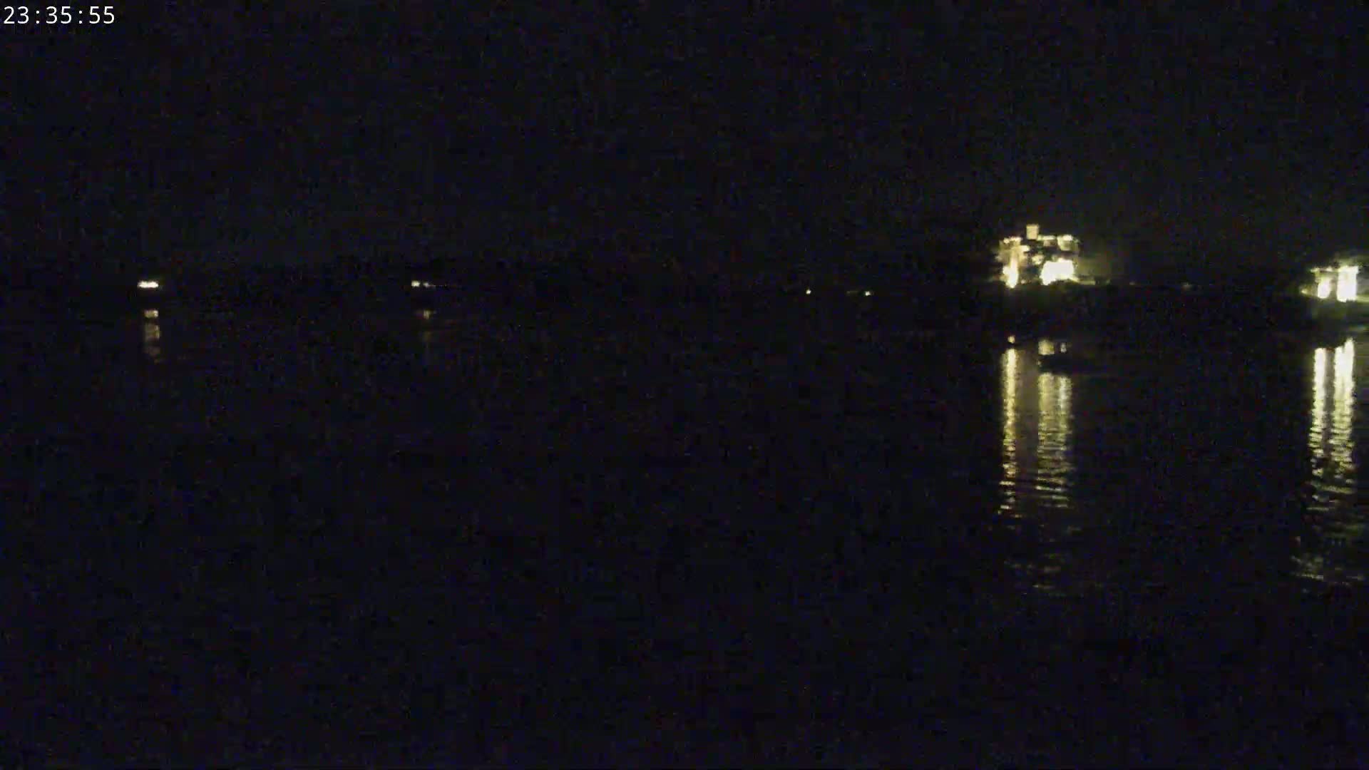 A dark nighttime scene shows a body of still water reflecting the lights of buildings on the far shore.