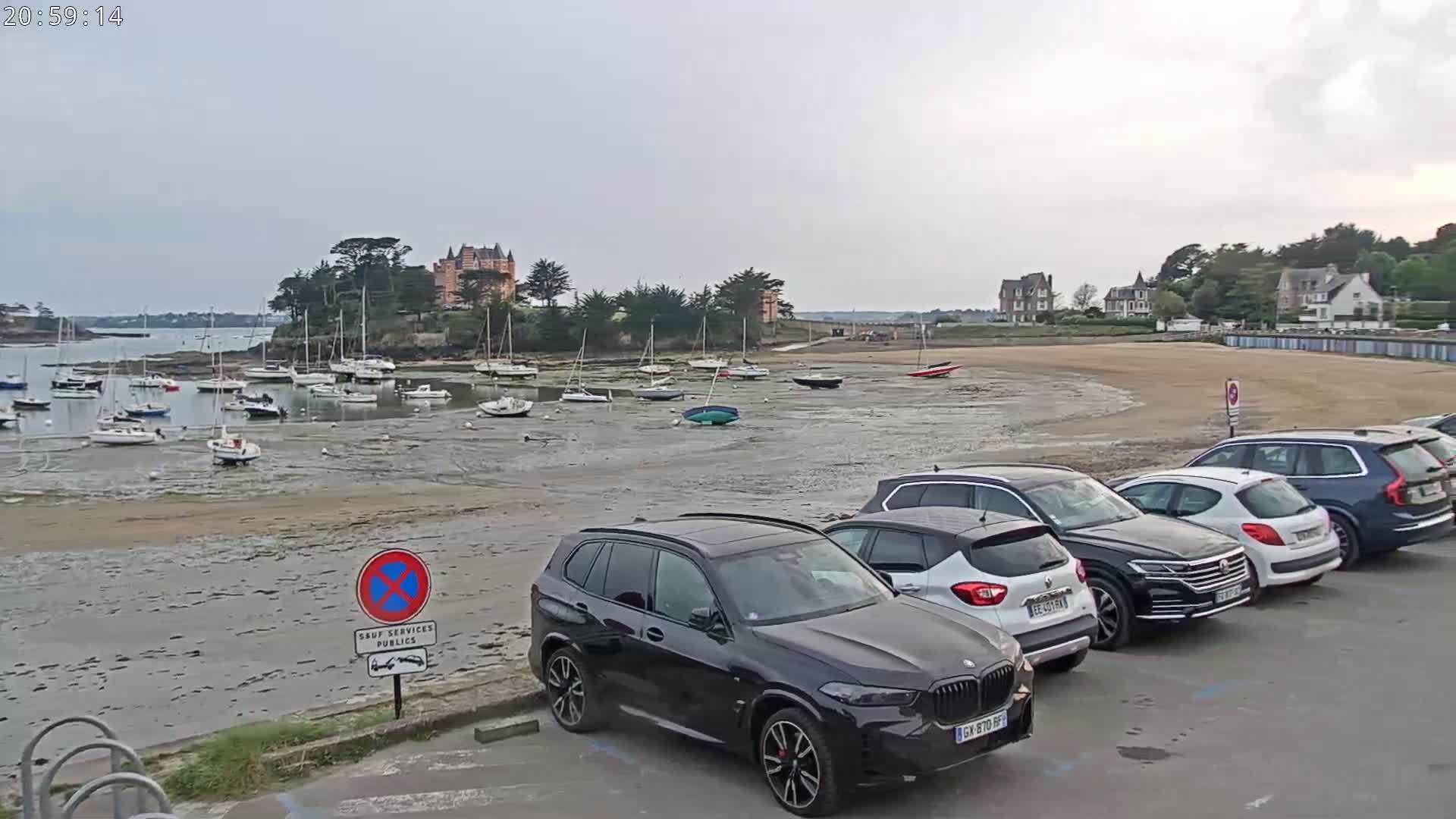 Many sailboats are moored in a calm bay at low tide, with several cars parked in a lot next to a sandy beach and buildings in the background.