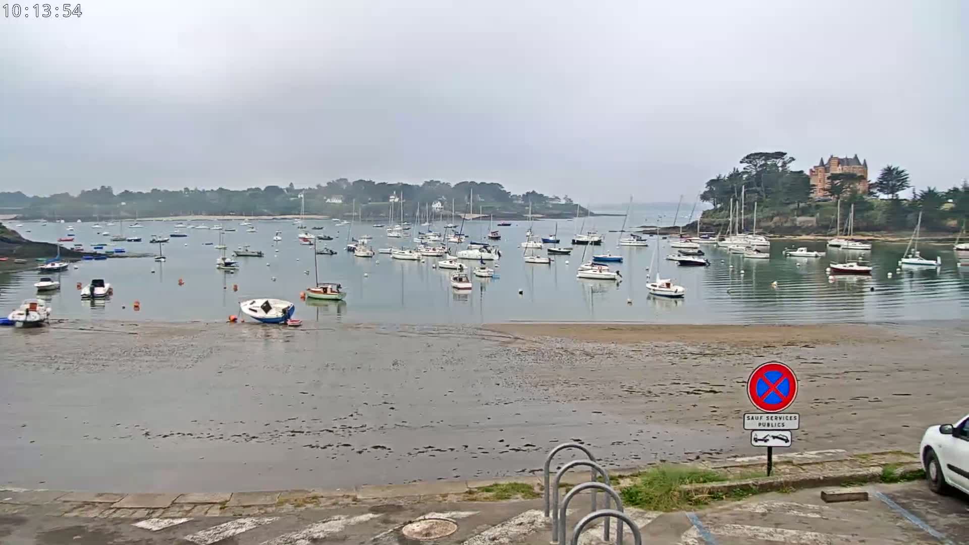 A calm, overcast day reveals a harbor filled with numerous sailboats at low tide, with a small sandy beach in the foreground and a building on a nearby hill.