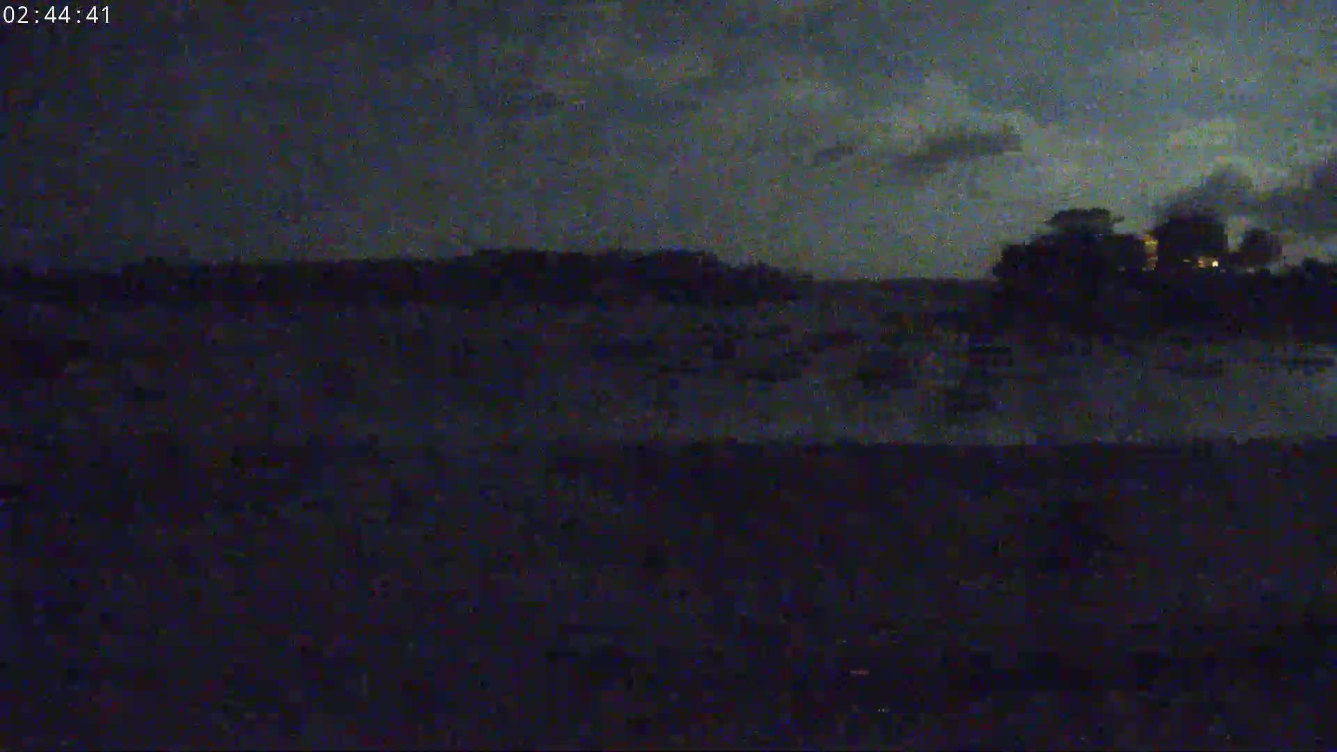 A dark, nighttime landscape shows a distant, dimly lit shoreline under a cloudy sky.