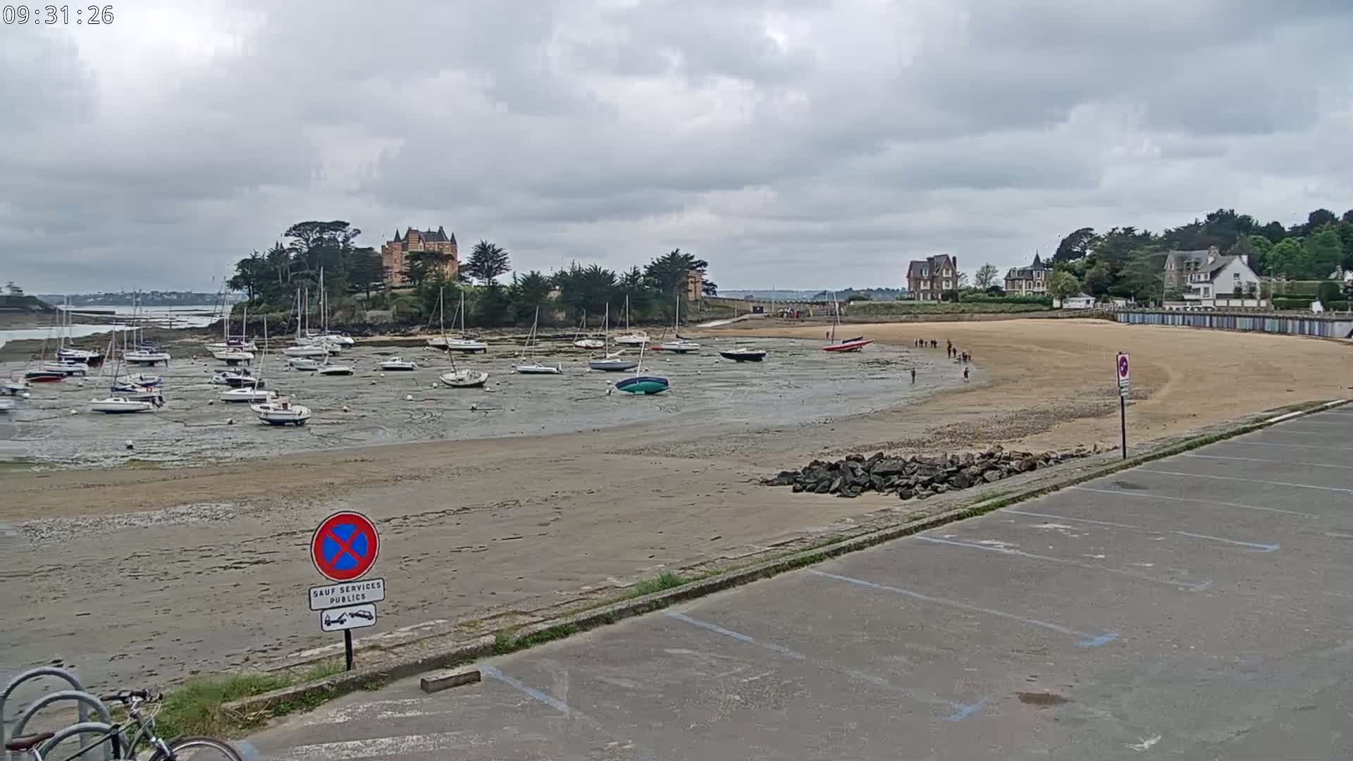 A sandy beach with several sailboats grounded at low tide near a waterfront with buildings under a cloudy sky.