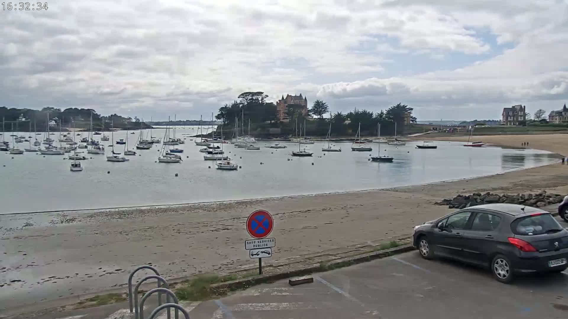 A calm bay with numerous sailboats at anchor under a cloudy sky, a sandy beach, and a dark-colored car parked nearby.