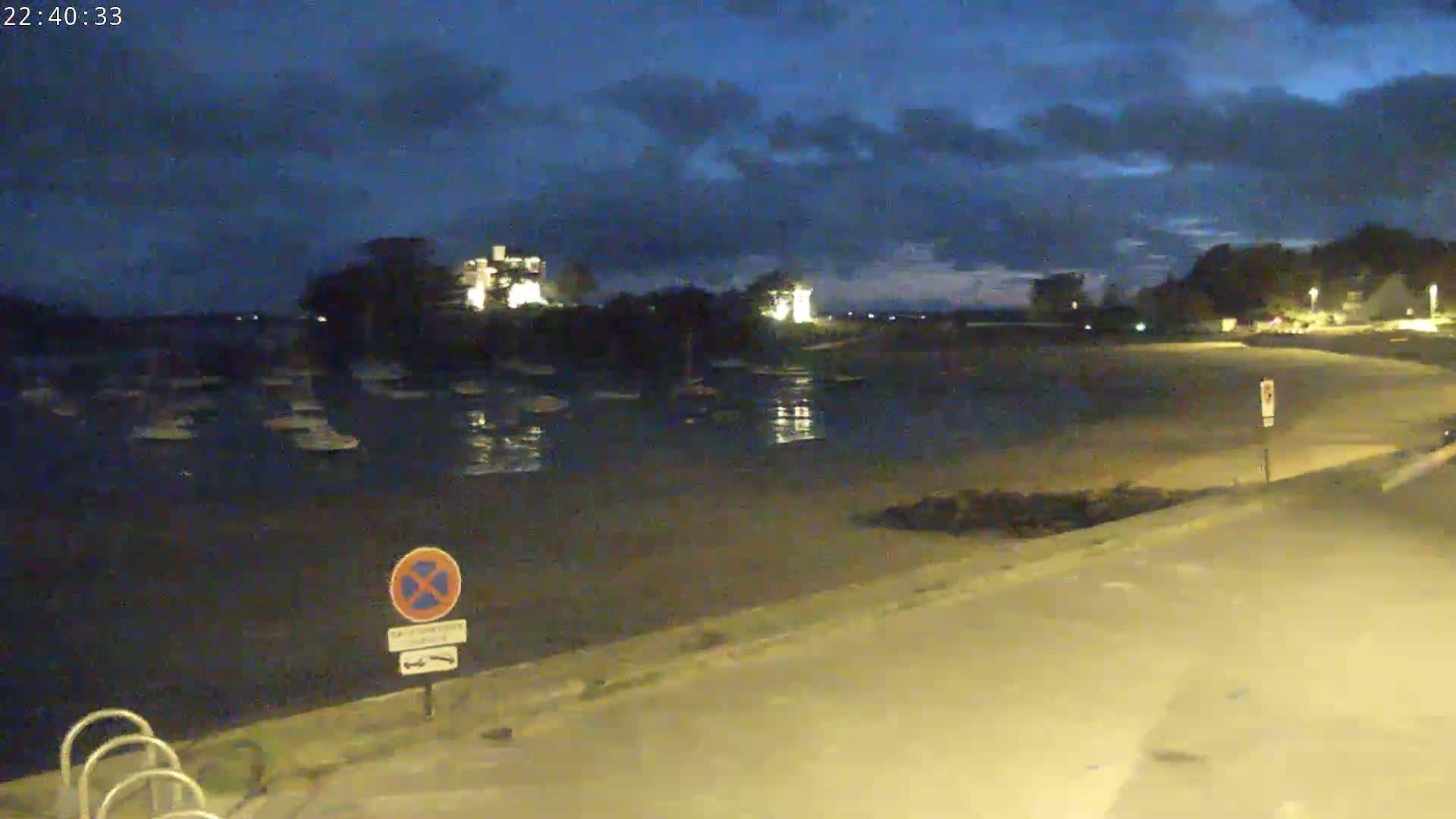 A nighttime view of a calm body of water with numerous boats moored, a shoreline with a paved path, and buildings illuminated in the distance under a cloudy sky.