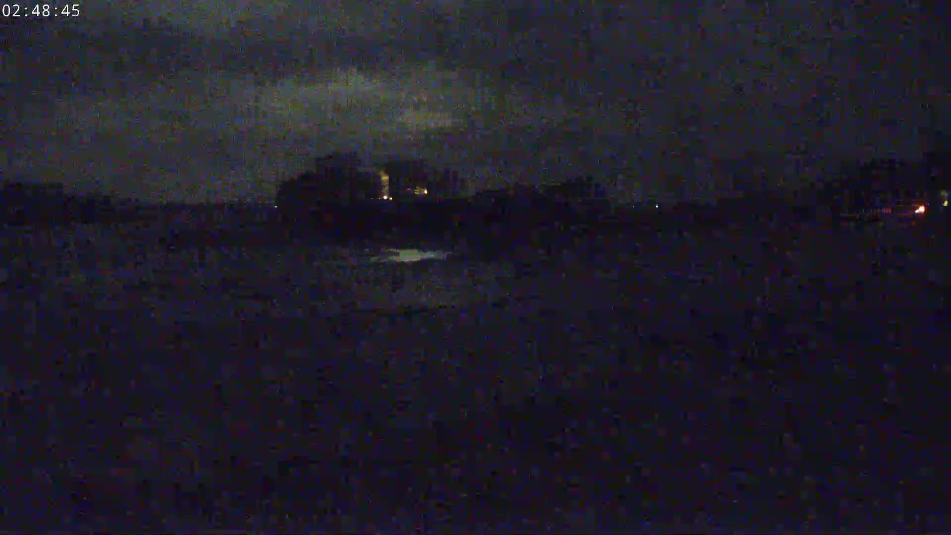 A dark, nighttime scene shows a distant, dimly lit landscape under a cloudy sky.
