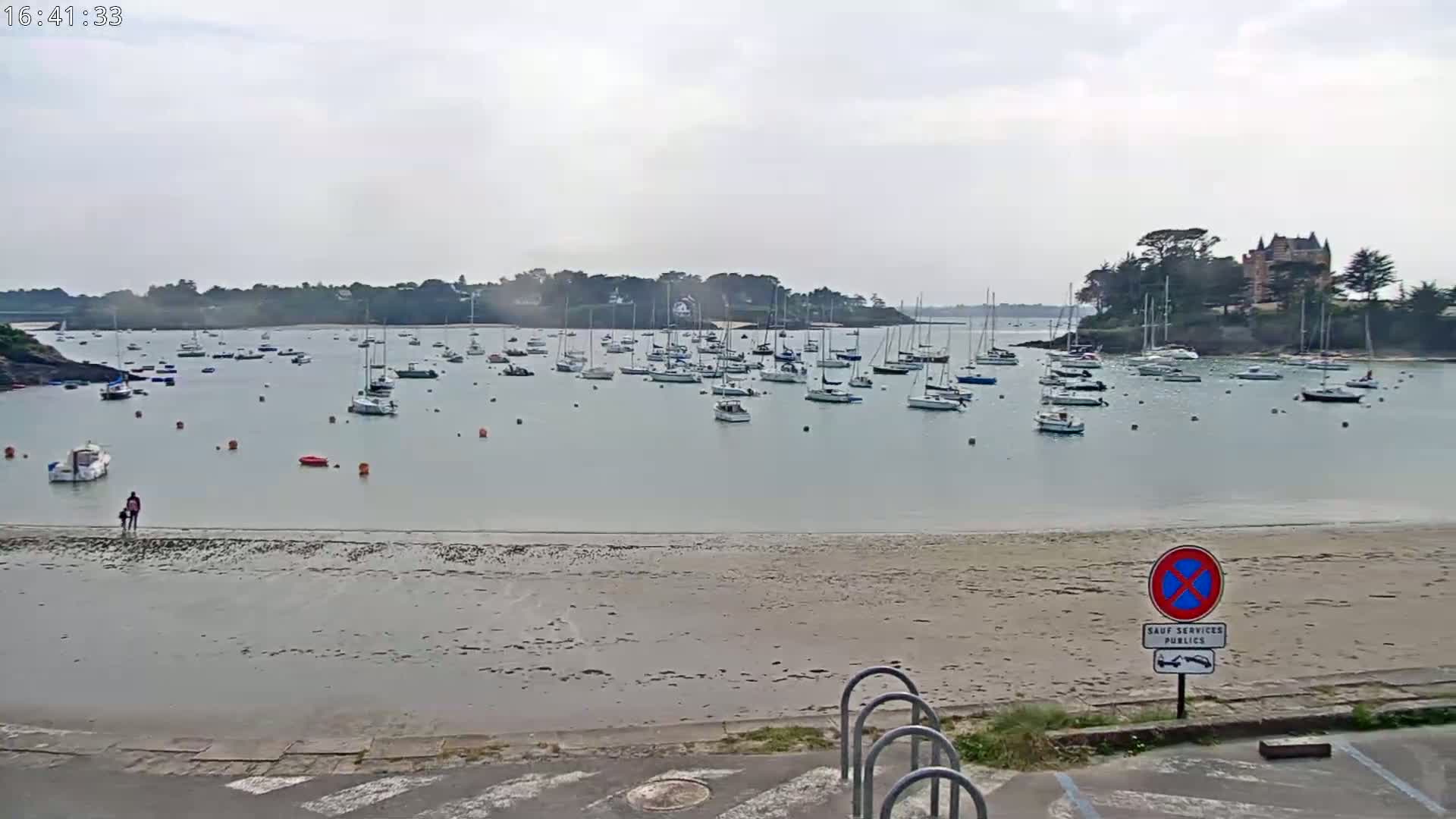 A calm bay with numerous sailboats at anchor is viewed from a sandy beach under an overcast sky.