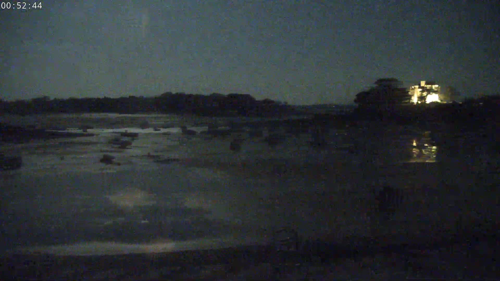 A dark, nighttime scene shows a body of water reflecting the lights of a distant building, with a dark shoreline and trees in the background.
