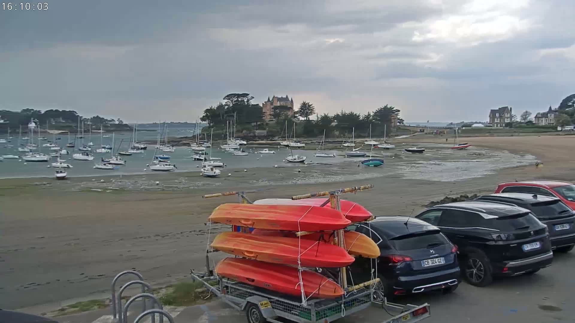 Kayaks are loaded onto a trailer parked on a sandy beach near a harbor full of sailboats under an overcast sky.