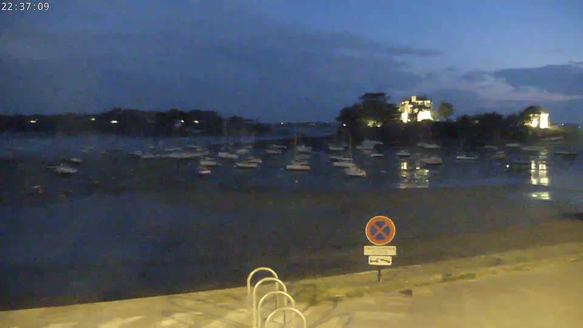 Many boats are moored in a calm body of water at dusk, with land and buildings visible in the background.