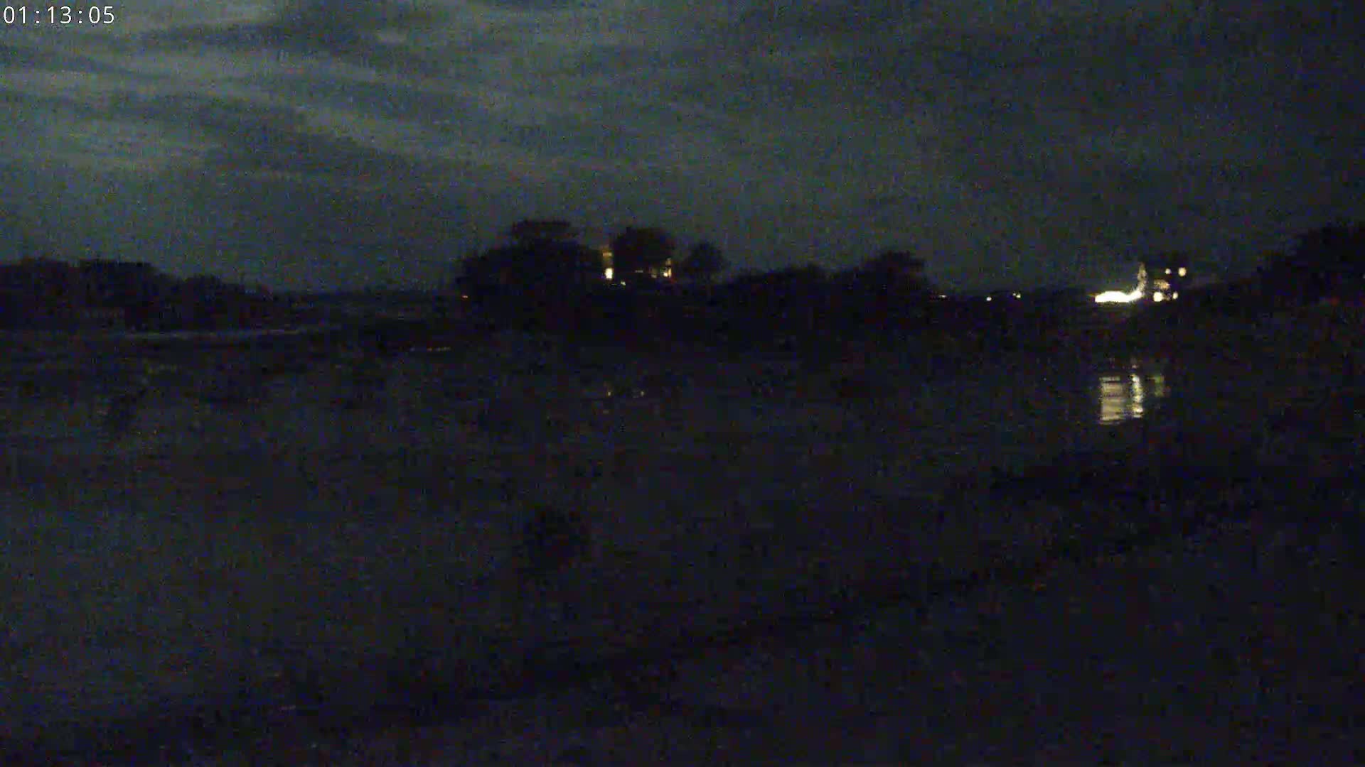 A dark, nighttime scene shows a body of water reflecting a few lights from buildings in the distance under a cloudy sky.