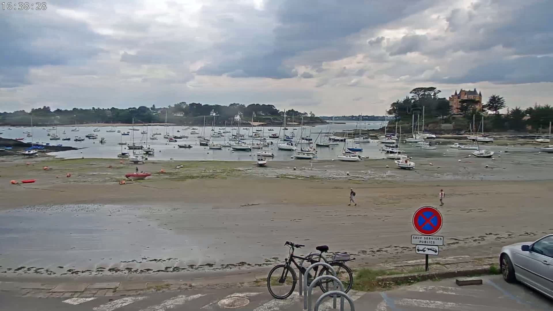A mostly sunny day shows a sandy beach with numerous sailboats in the water at low tide, a bicycle leaning against a bike rack, and two people walking on the beach.