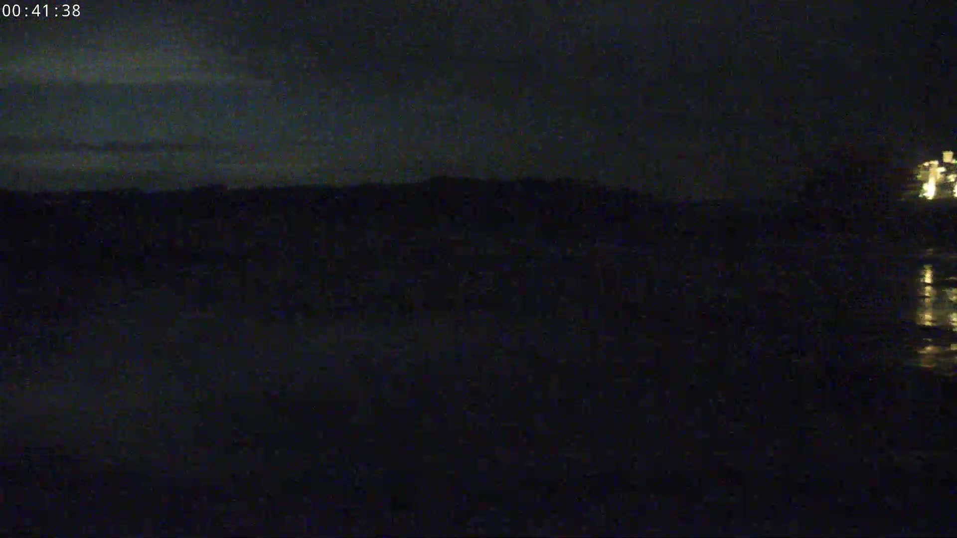 A dark, nighttime scene shows a body of still water reflecting a faint light source in the distance, with a dark landmass visible on the horizon under a mostly clear sky.
