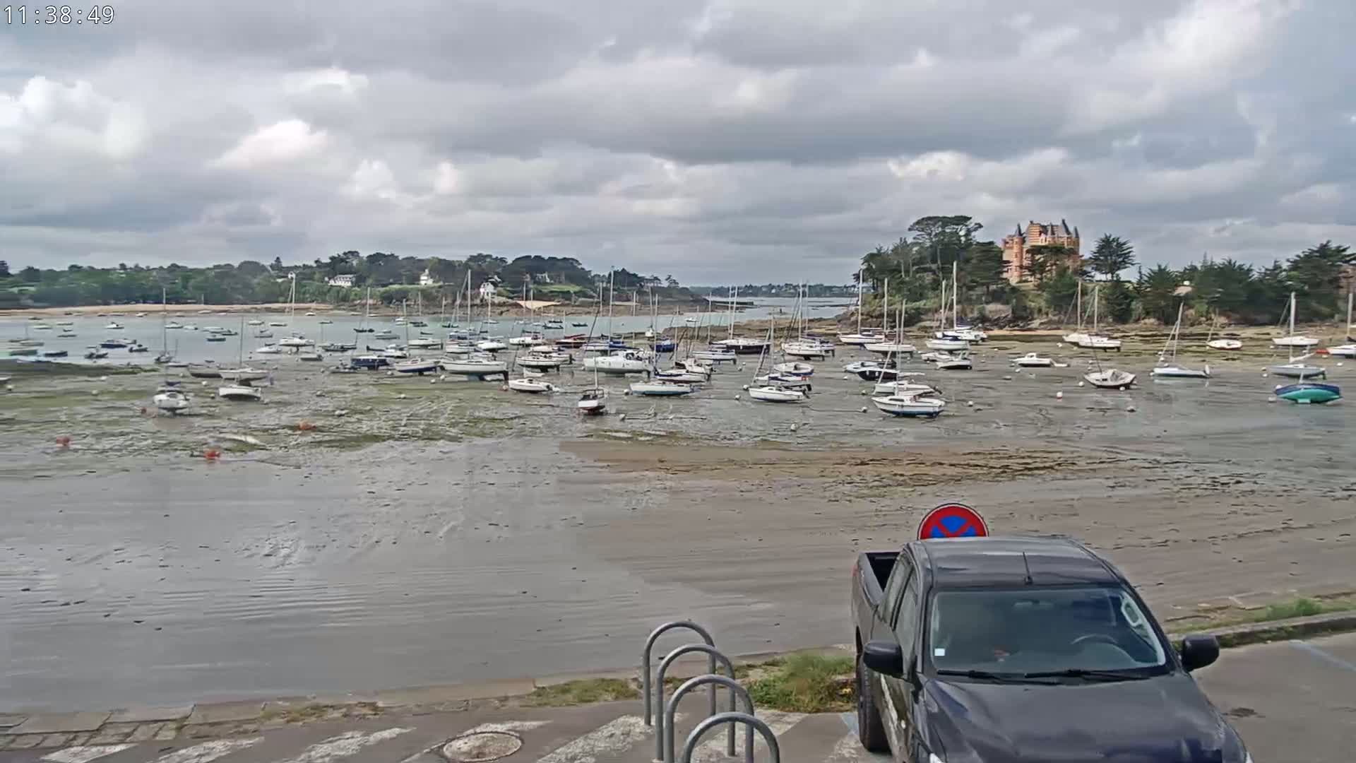 Many sailboats are grounded on a tidal flat in a harbor near a large house, under a cloudy sky.