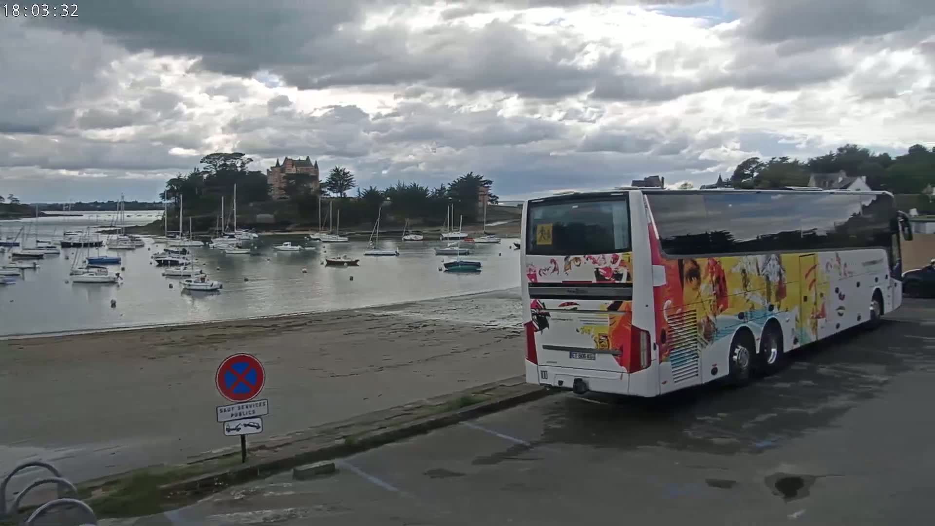 A colorful tour bus is parked near a sandy beach overlooking a calm harbor filled with numerous sailboats and a castle on a hill in the background under a cloudy sky.