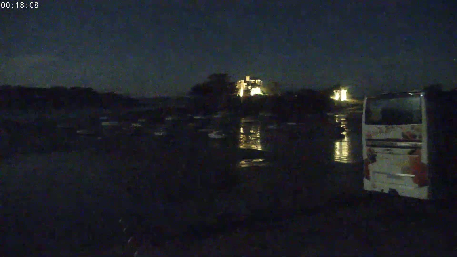 At night, a bus is parked near a body of water reflecting the lights of a building in the distance.