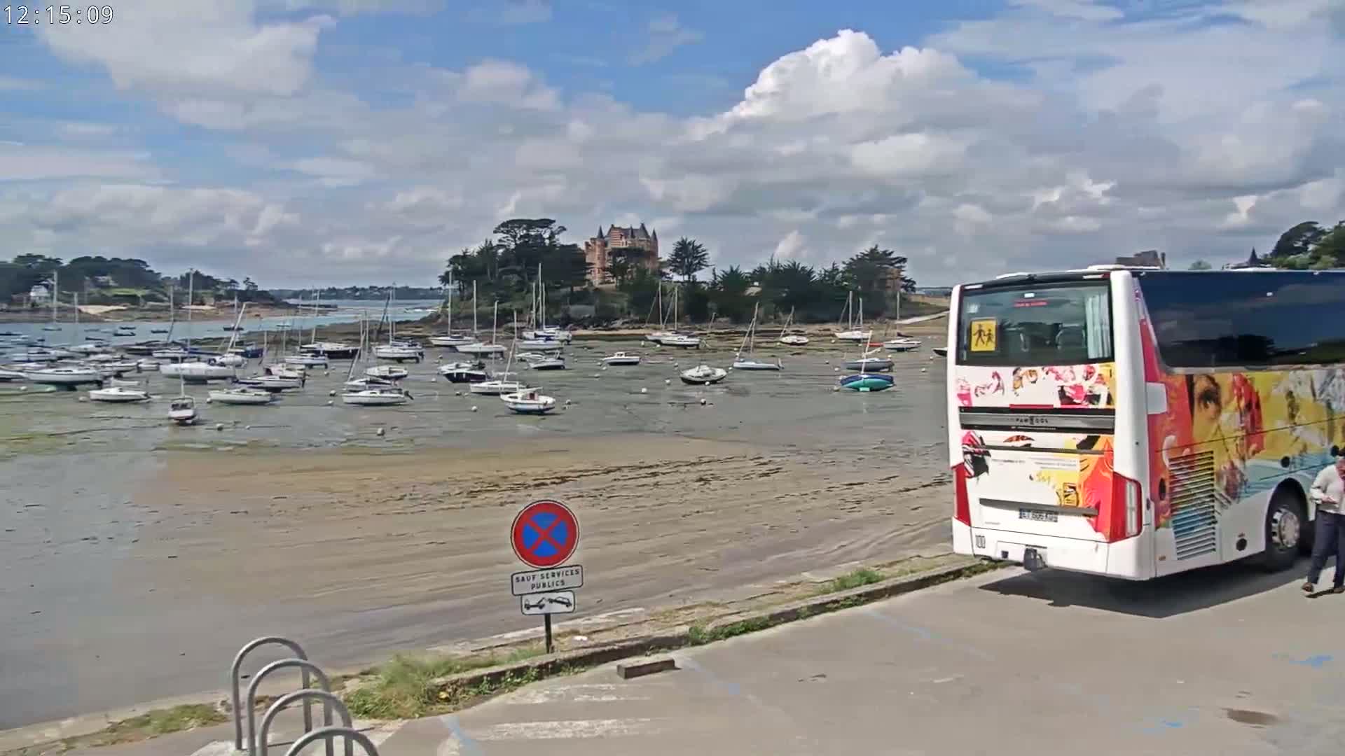 A partly sunny day shows a harbor filled with sailboats at low tide, a large bus parked nearby, and a chateau on a small island in the background.