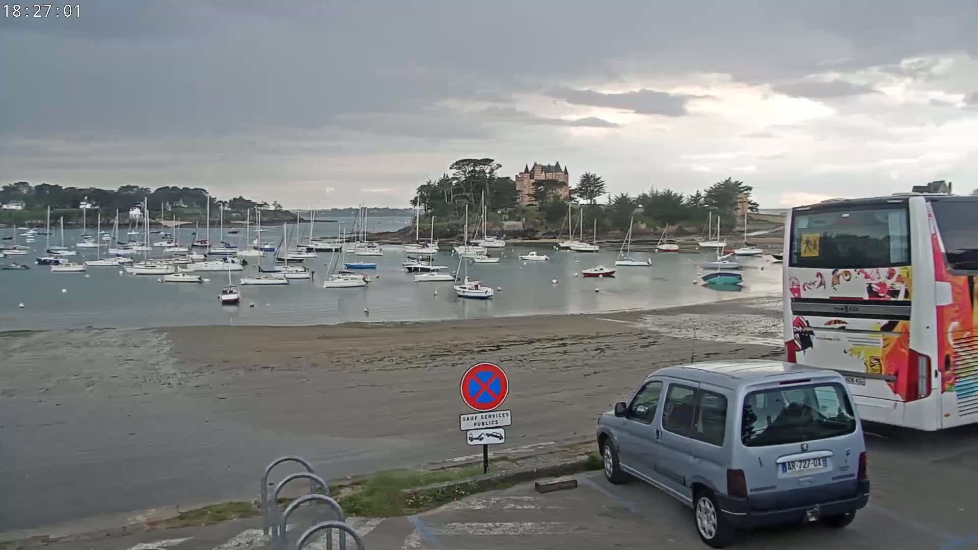 A harbor filled with sailboats under a cloudy sky is viewed from a sandy shore where a minivan and a bus are parked.
