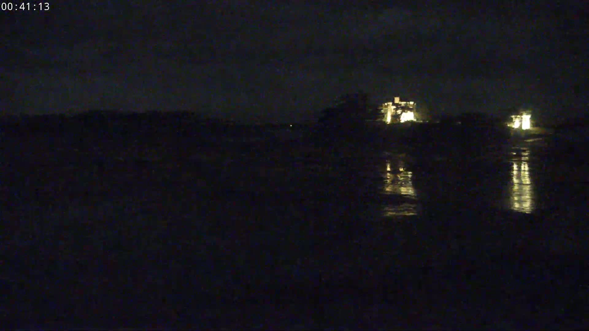A dark, nighttime scene shows a body of water reflecting the lights of buildings in the distance under a mostly clear sky.