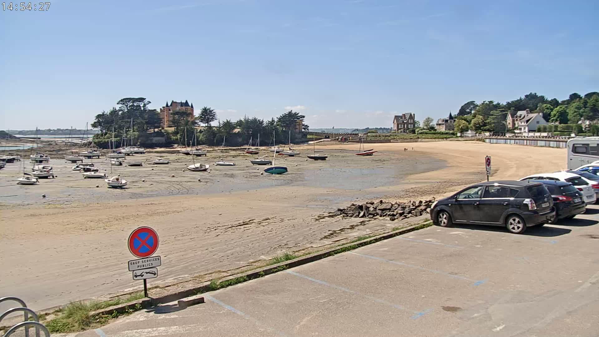 A sandy beach with numerous sailboats at low tide, parked cars, and buildings in the background under a clear, sunny sky.