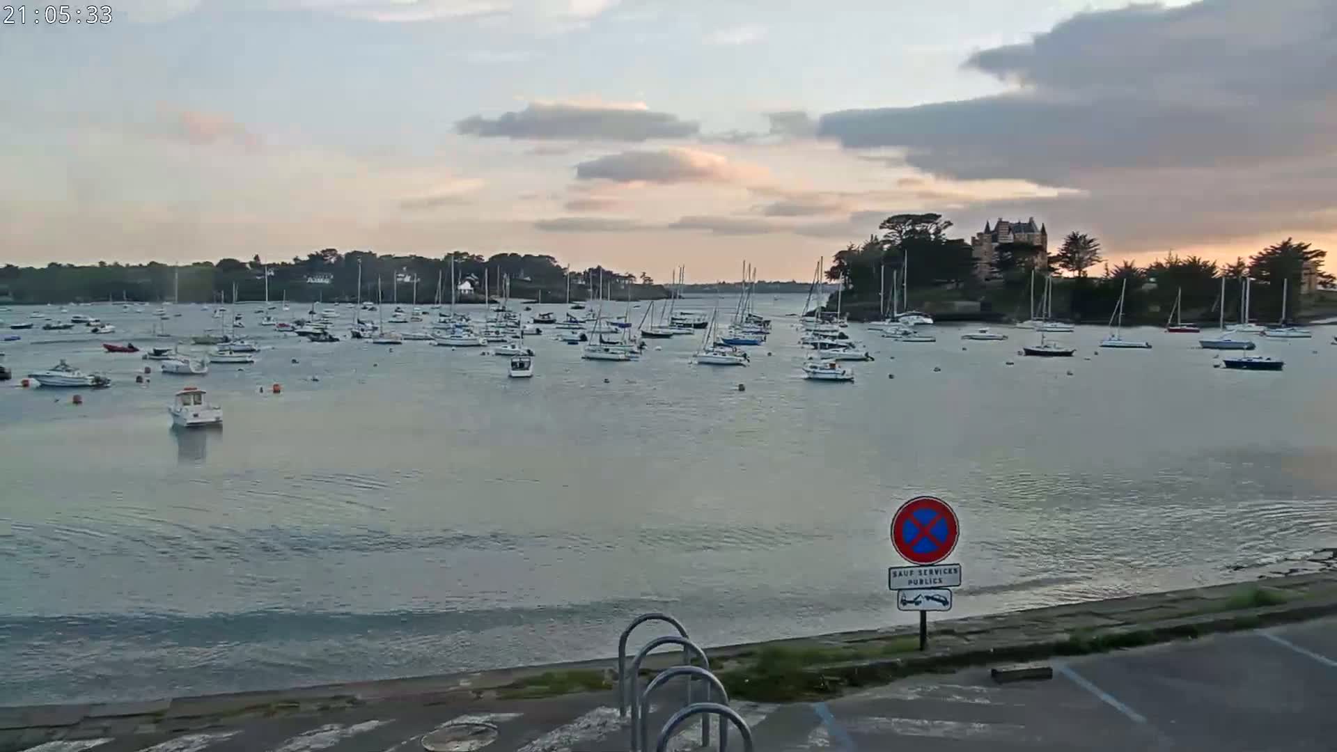 Many sailboats are moored in a calm bay under a partly cloudy evening sky, with a chateau visible on the far shore.