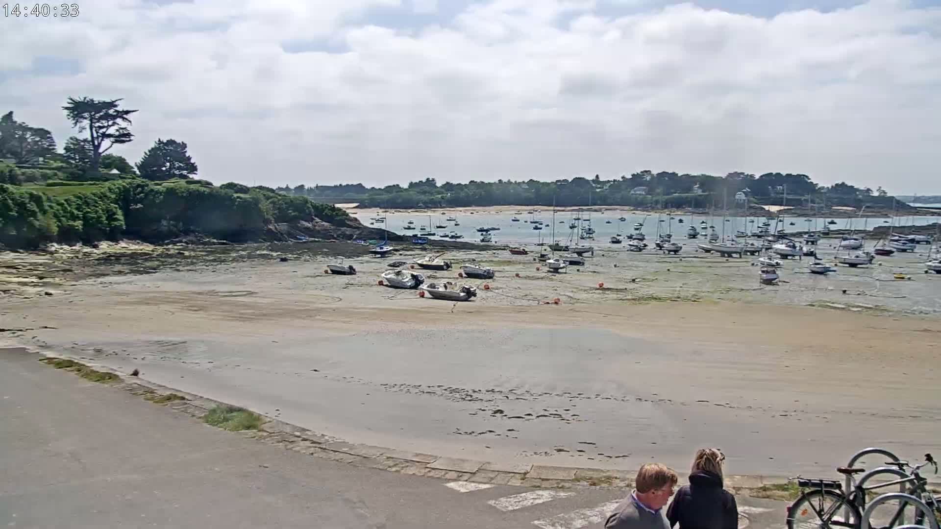 A sandy beach with numerous boats at low tide under a partly cloudy sky, with two people standing near parked bicycles in the foreground.