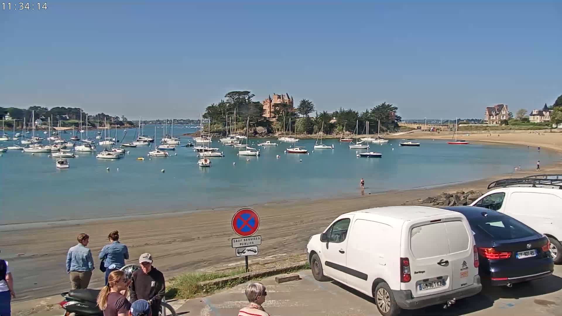 A sandy beach with numerous sailboats in the harbor, a castle on a small island in the background, and several people standing near parked vans on a sunny day.