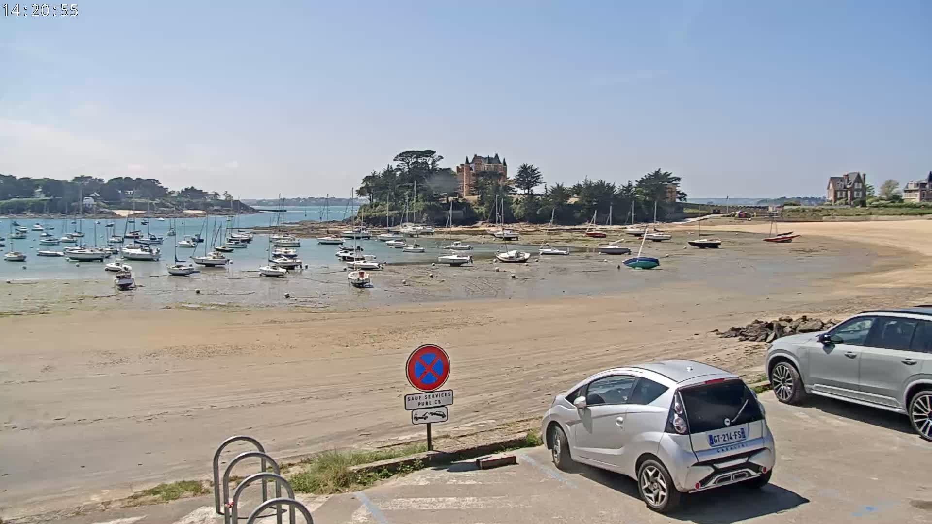 A sandy beach with numerous sailboats at low tide, a chateau visible in the distance, and two cars parked nearby under sunny skies.
